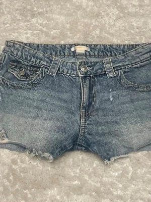Blå jeansshorts med rå kant - Snygga blå jeansshorts med rå, fransig kant och klassiska fem fickor. Shortsen har bälteshällor, knappgylf och slitna detaljer för en avslappnad look. Perfekta för varma dagar och enkel att matcha med olika toppar.