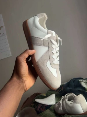 Vita och beige sneakers med mockadetaljer - Stilrena sneakers i vitt och beige med ovandel i slätt läder och detaljer i mocka. Skorna har vita snören och en klassisk gummisula i brunt. Perfekta för dig som gillar minimalistisk och clean stil.