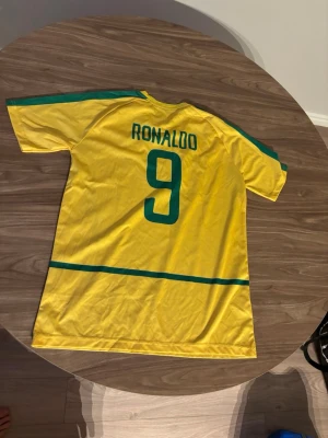 Gul Brasilien Ronaldo 9 matchtröja - Säljer en ikonisk gul Brasilien fotbollströja från Nike med gröna detaljer och nummer 9 samt Ronaldo på ryggen. Tröjan har officiellt Brasilien-emblem på bröstet och är tillverkad i lätt, ventilerande material. Perfekt för dig som älskar fotboll och klassiska landslagströjor.