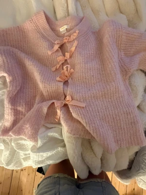 Ljusrosa kofta med rosetter Gina Tricot - Supersöt ljusrosa stickad kofta från Gina Tricot med fyra dekorativa rosetter framtill. Koftan har ribbstickade muddar och är gjord i ett mjukt, fluffigt material. Perfekt för dig som gillar gulliga detaljer och pastellfärger.