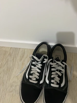 Svarta Vans Old Skool sneakers - Klassiska svarta Vans Old Skool sneakers med vit sula och vita snören. Skorna har den ikoniska vita sidolinjen och kontrastsömmar. Ovandelen är i canvas och mocka för en tidlös streetstil.