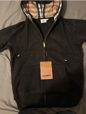 Svart Burberry hoodie med dragkedja - Svart hoodie från Burberry med hel dragkedja och klassiskt rutigt foder i huvan. Stora fickor framtill och justerbara snören i huvan med gulddetaljer. Perfekt för en stilren och lyxig streetwear-look.