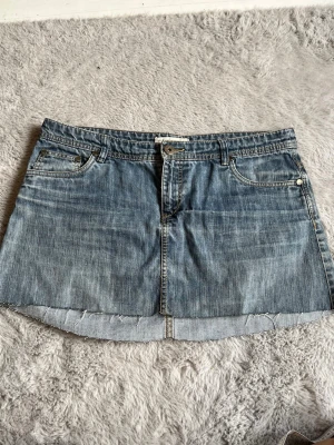 Kort jeanskjol  - Säljer en klassisk blå jeanskjol i minilängd med rå, avklippt kant. Kjolen har fem fickor, bälteshällor och knappstängning framtill. Perfekt till sneakers eller boots för en avslappnad look. Midjemått: 44cm Längd bak: 36cm Längd fram: 30cm