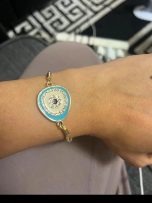 Guldarmband med blått öga och pärlor - Snyggt guldarmband med ett hänge i form av ett blått öga, dekorerat med små glittrande pärlor runt motivet. Armbandet har justerbar kedja och en modern, trendig design som sticker ut. Perfekt accessoar för dig som gillar unika detaljer och färg.