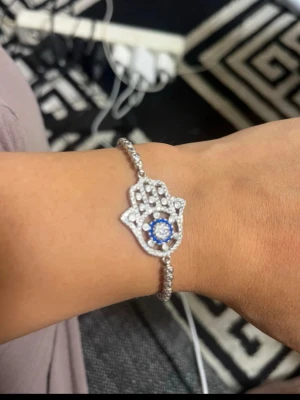 Silverfärgat armband med hamsa och pärlor - Stilrent armband i silverfärg med hamsa-hand dekorerad med små vita pärlor och en blå detalj i mitten. Armbandet har en tunn kedja med infattade pärlor längs hela bandet. Perfekt för dig som gillar unika smycken med symbolik.