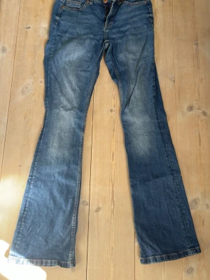 Blå bootcut jeans från Denim 1953 - Säljer ett par blå bootcut jeans från Denim 1953 i storlek 36. Jeansen har klassisk femficksmodell, slitningar och ljusare partier framtill för en cool vintagekänsla. Tillverkade i slitstark denim med snygga kontrastsömmar.