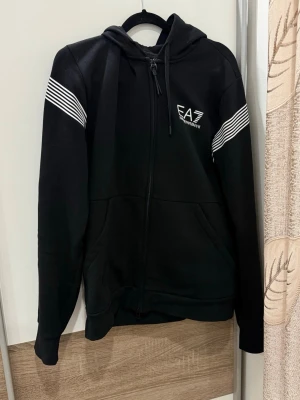 Svart hoodie från EA7 Emporio Armani - Snygg svart hoodie med dragkedja från EA7 Emporio Armani. Har vita ränder på ärmarna och logga på bröstet. Utrustad med huva och dragsko. Perfekt för en sportig och avslappnad stil.