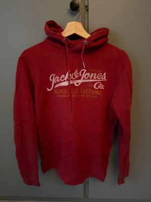 Röd hoodie från Jack & Jones - Snygg röd hoodie från Jack & Jones med tryck på bröstet där det står 'Rugged Denim'. Hoodien har dragsko i huvan och långa ärmar. Perfekt för en avslappnad och cool stil. Tillverkad i mjukt material för extra komfort.