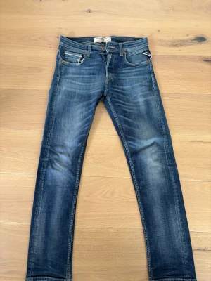 Blå slim fit jeans från Replay modell grover - Snygga blå slim fit jeans från Replay med klassisk femficksmodell och slitna detaljer. Jeansen har en tvättad look med ljusare partier på lår och knän samt Replay-logga på bakfickan. Tillverkade i denim med raka ben och normal midja. 27/30 storlek 