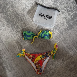 Färgglad bikini från Moschino - Supercool bikini från Moschino med bandeau-topp och knyt i mitten. Överdelen har grönt, blått och gult mönster, medan underdelen är röd med blommor och färgglada detaljer samt knyt i sidorna. Perfekt för sommarens strandhäng!