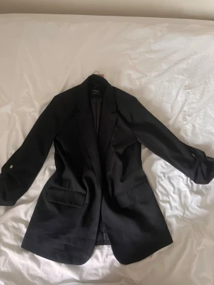 Svart oversized kavaj - Stilren svart oversized kavaj med klassisk krage och två framfickor. Kavajen har en rak passform och knäppning framtill. Perfekt att slänga över en enkel outfit för en cool look. Materialet ser ut att vara en blandning av polyester och viskos.