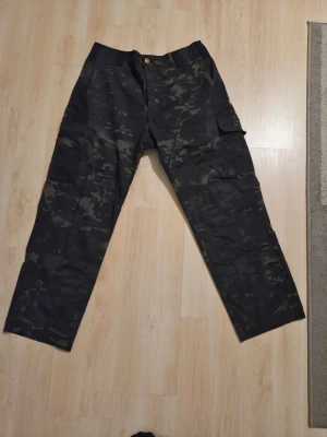 Cargobyxor Dark Camo - Säljer ett par svarta cargobyxor med mörkt camomönster. Byxorna har flera praktiska fickor på sidorna och är tillverkade i 100% bomull. Perfekta för dig som gillar streetwear och funktionella plagg. Byxorna är helt nya och aldrig använda. 