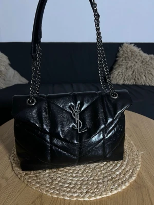 Svart quiltad handväska med kedjerem - Snygg svart quiltad handväska från Saint Laurent med silverfärgad YSL-logga framtill. Väskan har kedjeremmar i metall och är tillverkad i glansigt läder. Perfekt accessoar för att lyfta din outfit och rymmer det viktigaste du behöver ha med dig.