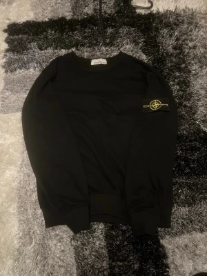 Svart sweatshirt från Stone Island - Svart sweatshirt från Stone Island med klassisk rund hals och ikonisk logotyp-patch på vänster ärm. Tröjan har ribbade muddar vid ärmslut och nederkant, vilket ger en snygg och avslappnad look. Perfekt för dig som gillar streetwear och stilrena plagg.