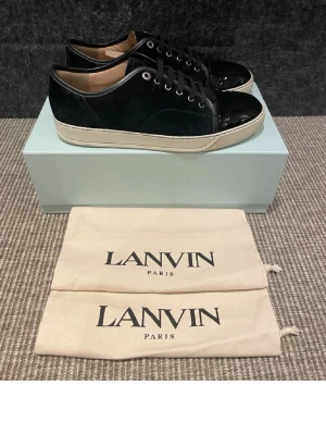 Svarta Lanvin - Snygga svarta sneakers från Lanvin med ovandel i mocka och lackdetaljer på tån. Skorna har vita sulor och svarta skosnören. Klassisk låg modell som passar till många olika stilar. Kommer med original dustbags.