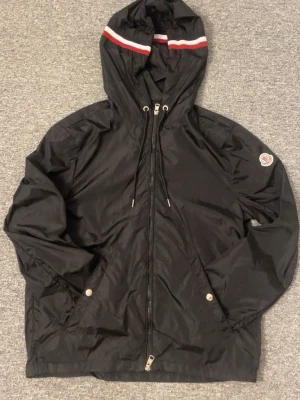 Svart Moncler vindjacka - Snygg svart vindjacka med huva och dragsko, samt vita och röda ränder på huvan. Jackan har hel dragkedja framtill, två fickor med tryckknappar och Moncler-logga på ärmen. Perfekt för blåsiga dagar och enkel att matcha till olika outfits.