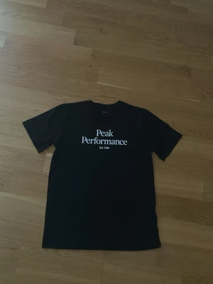Svart Peak Performance t-shirt - Snygg svart t-shirt från Peak Performance med vit logga och text på bröstet. Klassisk rund hals och korta ärmar. Tillverkad i mjuk bomull som känns skön mot huden. Perfekt till jeans eller shorts för en clean och enkel stil.