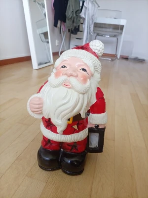 Dekorativ tomtelykta i keramik - Säljer en söt tomtefigur i keramik som fungerar som lykta. Tomten är röd och vit med svart bälte och skor, och har utskurna stjärnor på ryggen för ljusspridning. Perfekt som juldekoration eller mysig belysning i vintermörkret. Väll använt men funkar ändå 