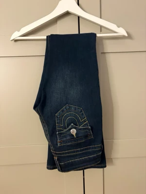 Mörkblå bootcut jeans med fickdetaljer - Snygga mörkblå bootcut jeans med kontrastsömmar och unika fickdetaljer bak. Jeansen har låg midja och silverfärgade knappar. Perfekta för dig som gillar en klassisk men ändå trendig look.