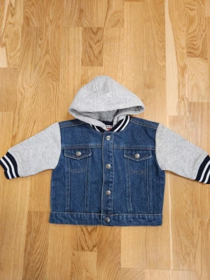 Bugs Bunny jeansjacka barn oanvänd - Snygg jeansjacka med gråa sweatshirtärmar och huva. Framsidan har tryckknappar och två bröstfickor. Baksidan pryds av en stor Bugs Bunny-tryck med texten 'what's up doc'. Storlek 74.