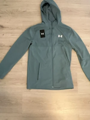Turkos hoodie med dragkedja från Under Armour - Snygg turkos hoodie från Under Armour med dragkedja framtill och huva. Logga på bröstet och stort Under Armour-tryck på ryggen. Perfekt för en sportig look och har praktiska fickor på sidorna.