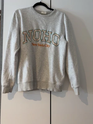 Grå sweatshirt med NOHO-tryck XS - Säljer en ljusgrå sweatshirt med texten NOHO New York City broderat framtill i grönt och orange. Tröjan har rund halsringning, ribbade muddar vid ärmslut och nederkant samt en avslappnad passform. Perfekt för en chill och trendig look.