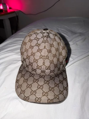 Gucci beige keps med GG-mönster - Säljer en beige keps från Gucci med det klassiska GG-monogrammönstret över hela kepsen. På sidan finns en grön och röd rand som ger extra Gucci-vibe. Justerbar rem baktill för perfekt passform. Materialet känns slitstarkt och exklusivt. sliten vid sidorna men ej synlig 