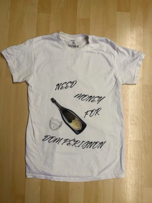 Vit t-shirt med Dom Pérignon-tryck - Vit t-shirt från Vonroe med tryck av en champagneflaska och texten 'Need money for Dom Pérignon' i svart. Klassisk passform och rund hals. Perfekt för dig som gillar statement-plagg och vill sticka ut.