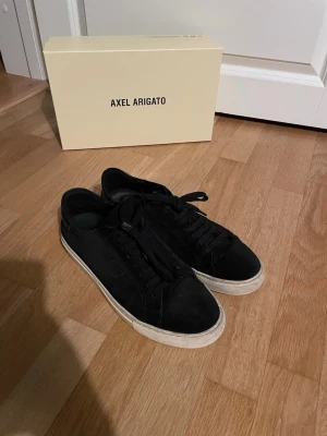 Svarta Axel Arigato sneakers - Svarta sneakers från Axel Arigato med klassisk låg profil och vit sula. Skorna är i mocka och har en stilren design med svarta skosnören. Perfekta för en clean och modern look. De är i bra begagnat skick. 