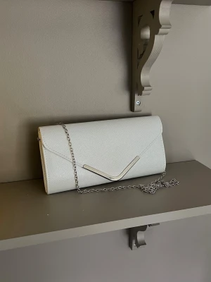 Silver kuvertväska med silverkedja - Stilren vit kuvertväska med elegant silverfärgad kedja och metallisk V-detalj på locket. Väskan har en slät, lätt skimrande yta och klassisk rektangulär form. Perfekt accessoar för dig som vill addera en clean och modern touch till din outfit. Nypris 400kr.