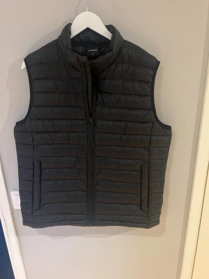Svart dunväst från Everest i nyskick! - Snygg svart dunväst från Everest med hög krage och dragkedja framtill. Västen har två sidofickor och quiltad design med horisontella sömmar. Perfekt för lager-på-lager och enkel att matcha med olika outfits.