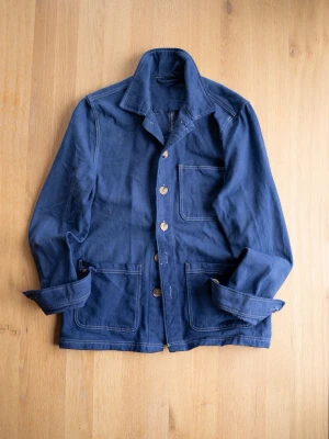 Blue Chore Jacket, size S - Stilren marinblå overshirt i bomull med tre stora fickor framtill och kontrasterande vita sömmar. Klassisk krage och knäppning med träknappar ger en avslappnad men snygg look. Perfekt att bära över en t-shirt eller hoodie.