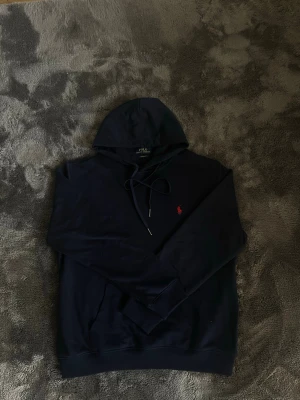 Mörkblå hoodie från Polo Ralph Lauren - Snygg mörkblå hoodie från Polo Ralph Lauren med klassisk huva och dragsko. På bröstet finns den ikoniska röda Polo-loggan broderad. Hoodien har långa ärmar, ribbade muddar och är tillverkad i mjukt material för maximal komfort.