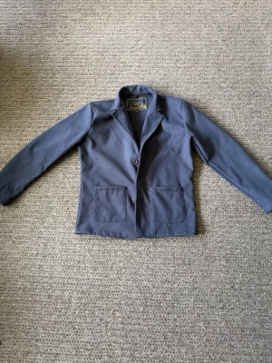 Mörkblå kavaj med två knappar - M/L. Stilren mörkblå kavaj med två knappar framtill och två stora fickor. Klassisk krage och rak passform. Materialet ser ut att vara bomull eller en bomullsblandning. Perfekt för en snygg och avslappnad look.