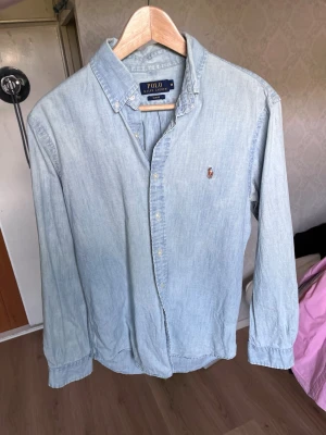 Ljusblå skjorta från Polo Ralph Lauren - Säljer en klassisk ljusblå skjorta från Polo Ralph Lauren med button-down krage och den ikoniska broderade loggan på bröstet. Skjortan har långa ärmar och knappar framtill. Perfekt till jeans eller chinos för en clean look.