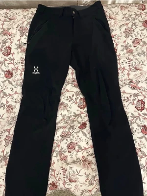 Svarta byxor från Haglöfs - Säljer ett par svarta byxor från Haglöfs med raka ben och dragkedjeförsedda fickor. Byxorna har en enkel design med logga på vänster lår och knappstängning i midjan. Perfekta för dig som gillar outdoor och funktionella plagg.
