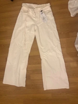 Vida vita jeans med knytband - Säljer ett par trendiga vita jeans med vida ben och knytband i midjan. Jeansen har hög midja och raka bakfickor. Perfekta för dig som gillar en avslappnad och modern stil.