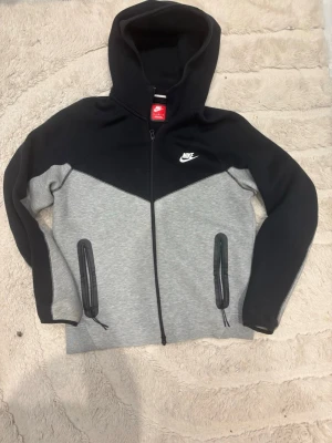 Nike tech  - Snygg hoodie från Nike i svart och grått med hel dragkedja framtill. Tröjan har huva, två fickor med dragkedja och Nike-logga på bröstet. Perfekt för en sportig och avslappnad stil. Materialet känns mjukt och bekvämt.