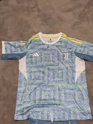Juventus träningsset Adidas blå - Juventus träningsset från Adidas med en blå och vit t-shirt med abstrakt mönster, vita och gula detaljer samt klubbmärke och Adidas-logga. Matchande blå shorts med vita och gula detaljer, elastisk midja och klubbmärke. Perfekt för fotbollsträning.