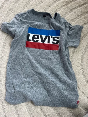 Grå Levi's t-shirt med logga - Snygg grå t-shirt från Levi's med den klassiska stora loggan i blått, vitt och rött framtill. T-shirten har korta ärmar och är gjord i mjuk bomull, perfekt för en avslappnad och trendig look. Storlek: 128-132