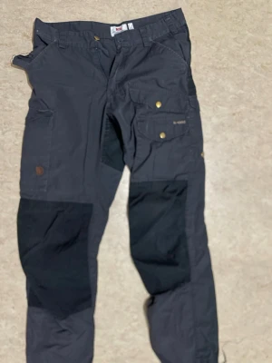Svarta fjällräven med förstärkningar - Snygga svarta cargobyxor med förstärkta partier på knän och bak, flera fickor med tryckknappar och detaljer i mässing. Tillverkade i slitstarkt material, perfekta för en aktiv livsstil. Klassisk cargomodell med avslappnad passform.