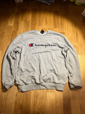 Grå Champion sweatshirt M - Snygg grå sweatshirt från Champion med klassisk logga broderad på bröstet. Tröjan har rund halsringning, ribbade muddar och är tillverkad i mjukt material som passar perfekt till en avslappnad stil.