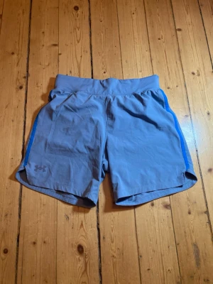 Ljusblå träningsshorts från Under Armour - Säljer ett par ljusblå träningsshorts från Under Armour med blåa detaljer längs sidorna. Shortsen har elastisk midja och är tillverkade i ett lätt och snabbtorkande material, perfekta för gymmet eller löprundan.