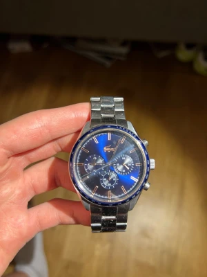 Lacoste klocka med blå urtavla - Stilren Lacoste klocka med blå urtavla, silverfärgat armband i metall och tre mindre urtavlor för kronograf. Klockan har tydliga indexmarkeringar och knappar på sidan för extra funktioner. Perfekt accessoar för dig som gillar klassisk design.