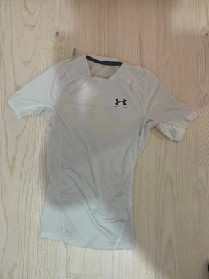 Vit kompressionströja Under Armour - Säljer en vit kompressionströja från Under Armour med korta ärmar och rund hals. Om Tröjan har logga på bröstet och ryggen, och är tillverkad i ett stretchigt funktionsmaterial som andas. Perfekt för träning eller sportaktiviteter.