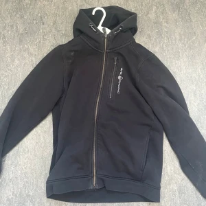 Svart hoodie från Sail Racing - Svart hoodie från Sail Racing med dragkedja framtill och en bröstficka med logga. Hoodien har huva med dragsko och ribbade muddar. St 160