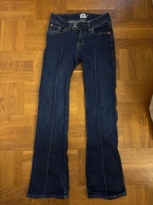 Blå low waist bootcut jeans - Säljer ett par blå low waist jeans med bootcut passform och dubbla knappar i midjan. Jeansen har dekorativa sömmar framtill och klassiska bakfickor. Perfekta för dig som gillar en retro vibe och vill ha ett par jeans som sticker ut lite extra.