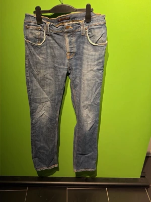 Blå nudie jeans - Säljer ett par klassiska blå jeans med raka ben och kontrastsömmar. Jeansen har fem fickor, bälteshällor och detaljer i orange söm. Materialet är denim i bomull och passformen är normal. Perfekta till en avslappnad stil. De har ett hål i höger ficka som visas på sista bilden men de borde gå o laga utan så mycket problem 😄😊