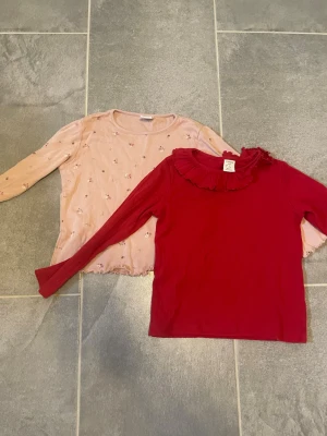2 långärmade tröjor  - Snygg röd långärmad topp i ribbat material med en volangdetalj runt halsen. Perfekt för dig som vill ha en enkel men ändå trendig look. Toppen har en figurnära passform och är mjuk och stretchig.