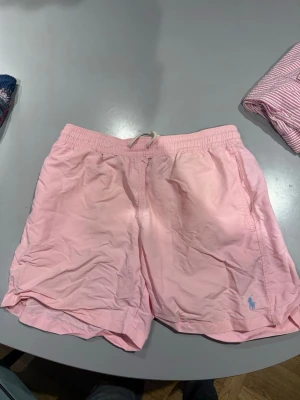 Ljusrosa Ralph Lauren badbyxor med logga - Säljer ett par ljusrosa badbyxor med elastisk midja och dragsko. De har en liten blå logga broderad framtill och en bakficka med knapp. Klassisk modell som passar perfekt till stranden eller poolen.